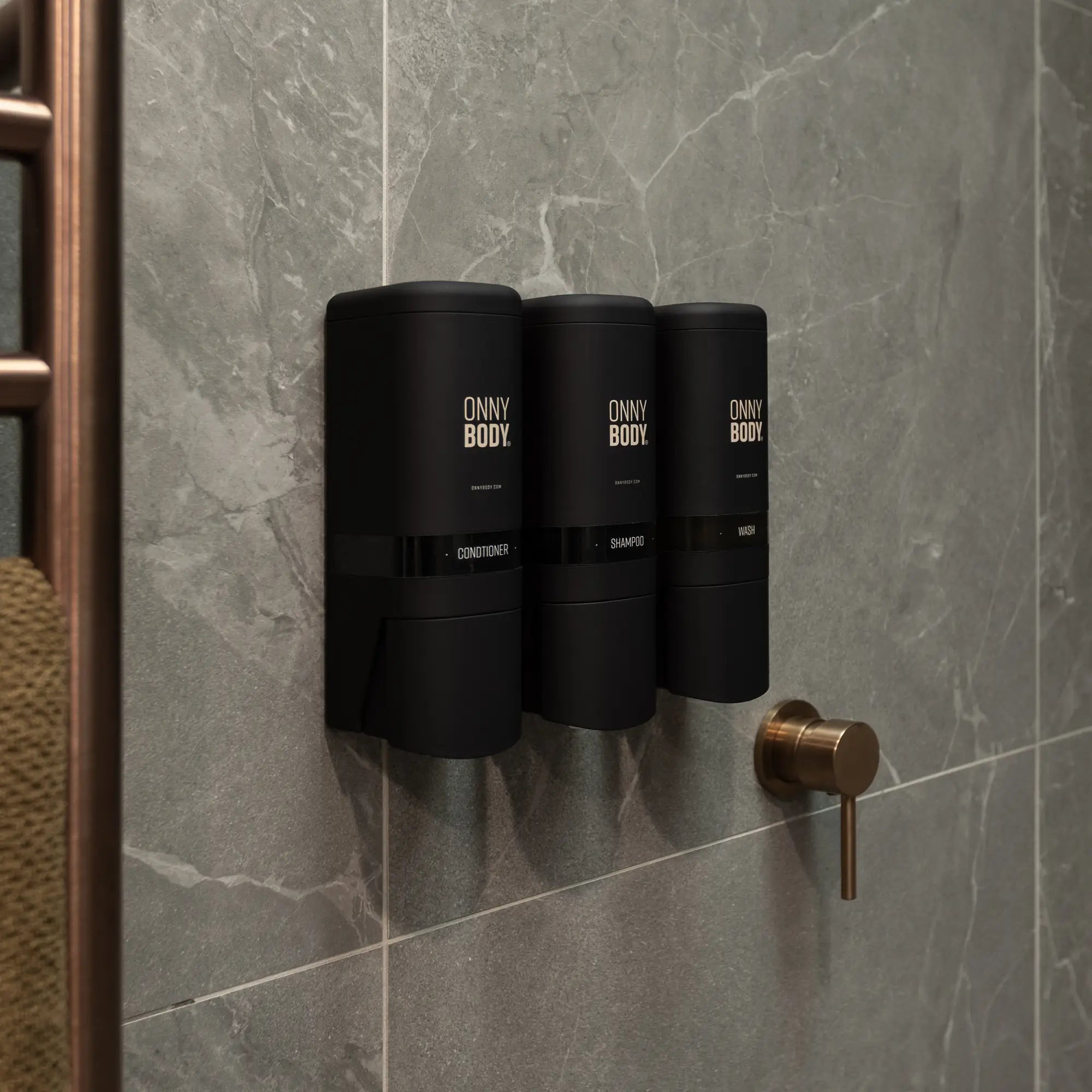 Three black ONNYBODY dispensers mounted on a grey tiled wall with a gold shower handle.